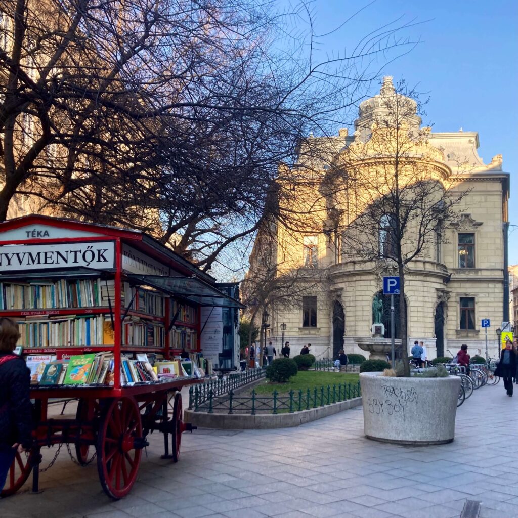 The picture stands for Workation and remote work in Budapest. On the left you can see a red book trolley and behind it on the right a two-storey baroque building, which is supposed to represent the Szabo Ervin Library.