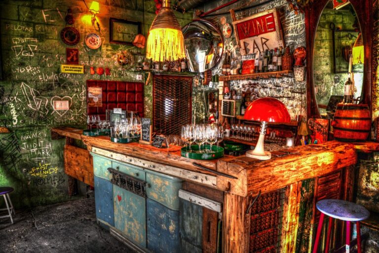 Das Bild steht für Ruin Bars Budapest. Man sieht im Zentrum eine bunte wild zusammengehämmerte Bartheke. Sie besteht aus mehreren Holzplatten und ihre Front ist eine blaue bunte Kommode. Auf der Bartheke stehen Weingläser, bereit um serviert zu werden. Ebenfalls steht rechts eine rote Tischlampe. Über der Bar hängt eine gelb leuchtende Deckenlampe mit langen Fransen. Hinter der Theke ist eine grüne Wand, welche komplett zugekritzelt ist mit weißen Filzstiften. Vereinzelt hängen zudem Spiegel, Teller und Bilderrahmen an der Wand. Man schaut rechts schräg auf die Bar.