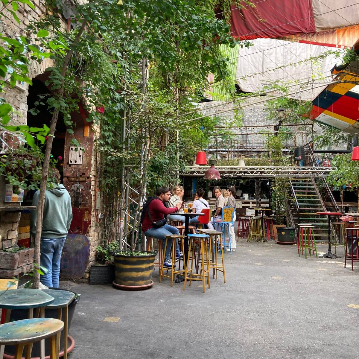 Auf dem Bild ist das Szimpla Kert zu sehen. genauer genommen der Innenhof. Viele Kletterpflanzen auf den wänden und bunt zusammegestellte Barhocker und Tische. im Hintergrund sieht man sogar eine Stiege die auf eine Galerie führt.