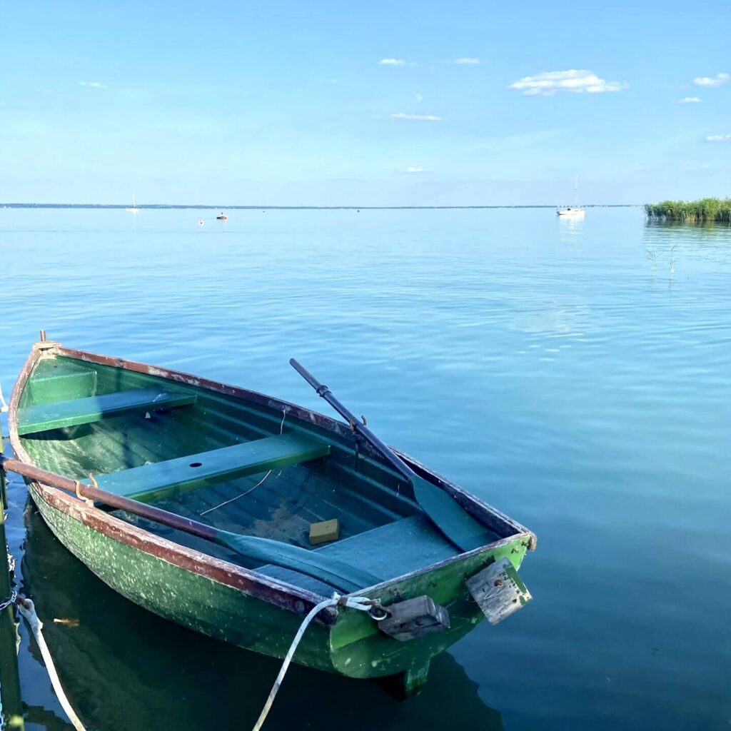 Das Bild steht symbolisch für Urlaub am Balaton. Man sieht im linken unteren Bildrand ein kleines grünes Holzboot, das auf einen beinahe leeren und stillen See schwimmt. Im rechts hinten sieht man bereits einen Teil von einem Schilfbewuchs und links davon treibt in der Ferne ein weißes Segelboot vorbei.
