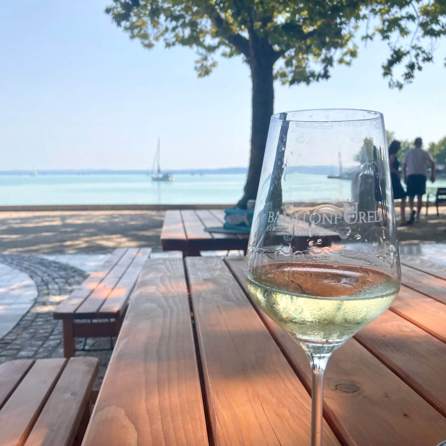 The image captures a serene and picturesque scene at Lake Balaton. In the foreground, a glass of white wine, branded with "Balatonfüred", rests on a wooden picnic table, inviting a moment of relaxation. The background reveals a calm, clear lake with a sailboat gently gliding on the water, adding to the tranquil atmosphere. A tree provides shade, while distant figures and more picnic tables suggest a pleasant social setting. The overall ambiance exudes peace and enjoyment, perfectly embodying the leisurely charm of a day spent at Lake Balaton.