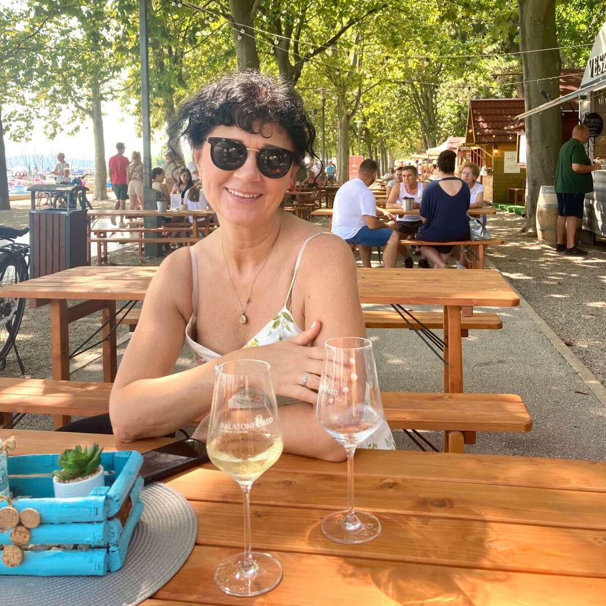 The picture shows my mum sitting at an outdoor table during the "Borhetek" (Wine Weeks) event in Balatonfüred, next to Lake Balaton. She is wearing sunglasses and a light, summery dress with thin straps, smiling warmly at the camera. On the table in front of her are two wine glasses, one containing white wine, and a decorative blue wooden box with a small potted plant. In the background, people are seated at other tables, enjoying the event under the shade of large trees. The overall atmosphere is relaxed and sunny, characteristic of a pleasant summer day.