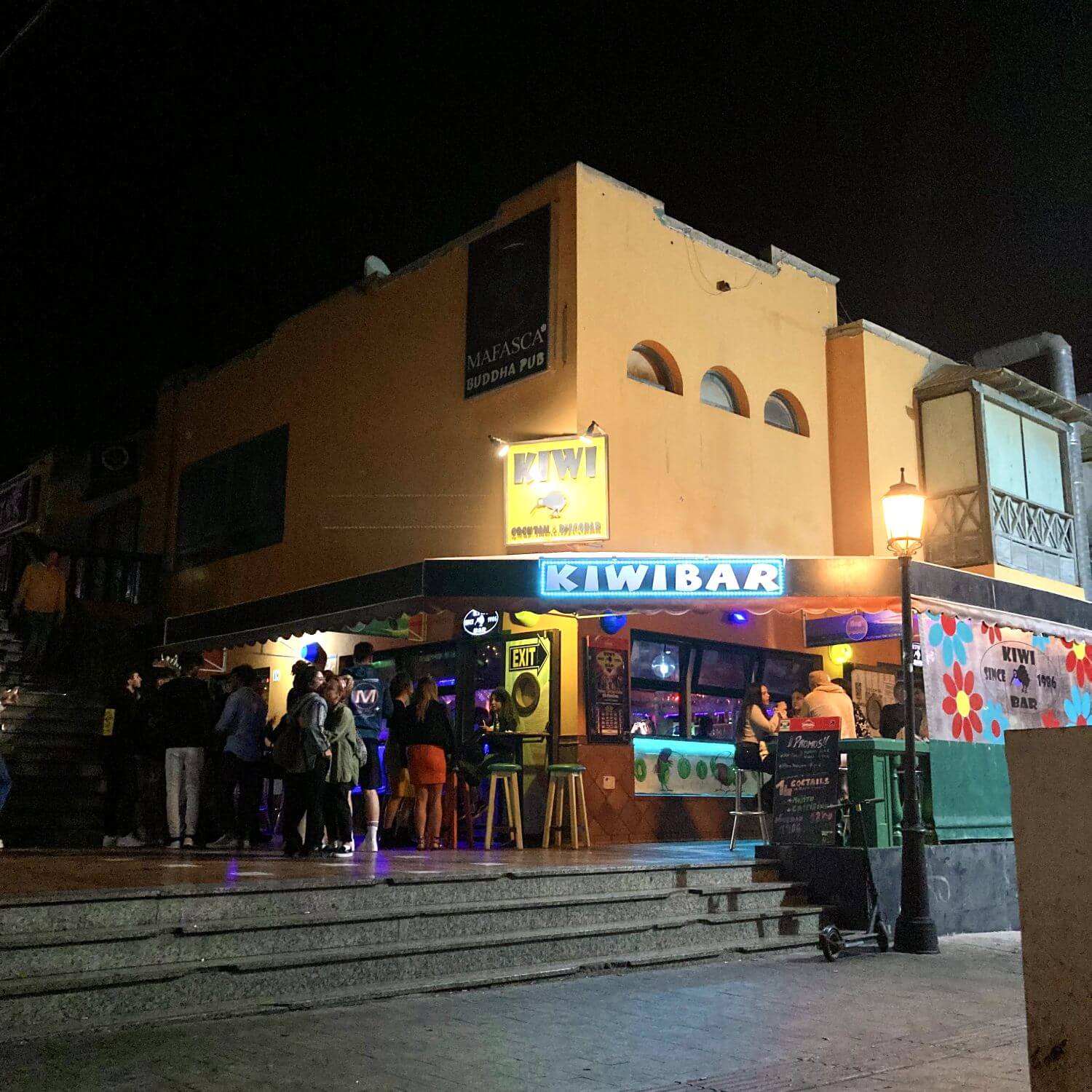 Das hochgeladene Bild zeigt die Kiwi Bar, Teil Corralejo Party Welt, die eine lebhafte "Fuerteventura Party"-Atmosphäre vermittelt. Die Bar befindet sich in einem orangefarbenen Gebäude und ist durch ein auffälliges, beleuchtetes Schild mit der Aufschrift "KIWI BAR" leicht erkennbar. Vor der Bar versammeln sich Menschen, die sich unterhalten und Getränke genießen. Die Bar selbst ist bunt beleuchtet und wirkt einladend. Im oberen Teil des Gebäudes ist ein weiteres Schild für den "Mafasca Buddha Pub" zu sehen. Die Szene strahlt eine lebendige und gesellige Stimmung aus, typisch für das Nachtleben in Corralejo.