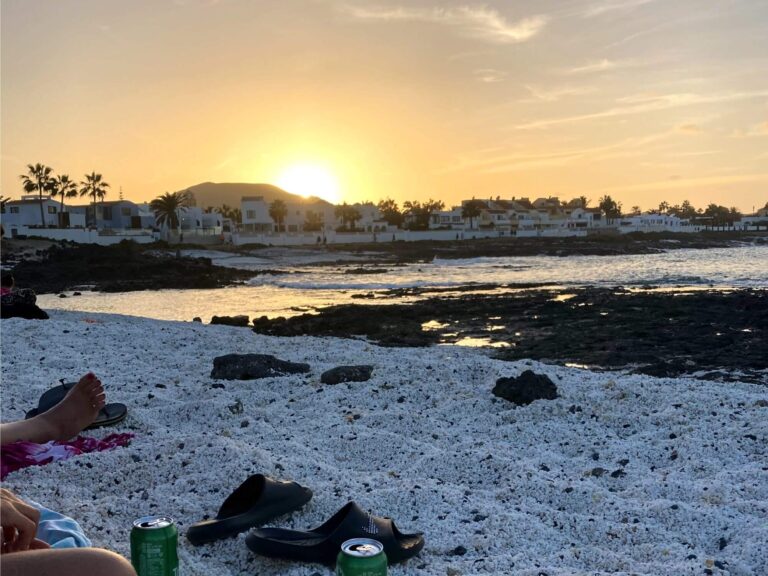 Das hochgeladene Bild zeigt einen friedlichen Strand bei Sonnenuntergang, der eine entspannte "Fuerteventura Party"-Atmosphäre vermittelt. Der Corralejo-Strand besteht aus hellem, grobkörnigem Sand oder Kieselsteinen. Im Vordergrund sind persönliche Gegenstände wie Sandalen und eine Getränkedose zu sehen, während Menschen am Strand entspannen. Felsige Bereiche führen ins ruhige Meer, das die goldenen Farben des Sonnenuntergangs reflektiert. Im Hintergrund stehen weiße Häuser einer Küstensiedlung, umgeben von Palmen. Die Sonne geht hinter den Häusern und einem Hügel am Horizont unter, der Himmel ist in goldene und orangefarbene Töne getaucht, was der Szene eine warme und gemütliche Atmosphäre verleiht.
