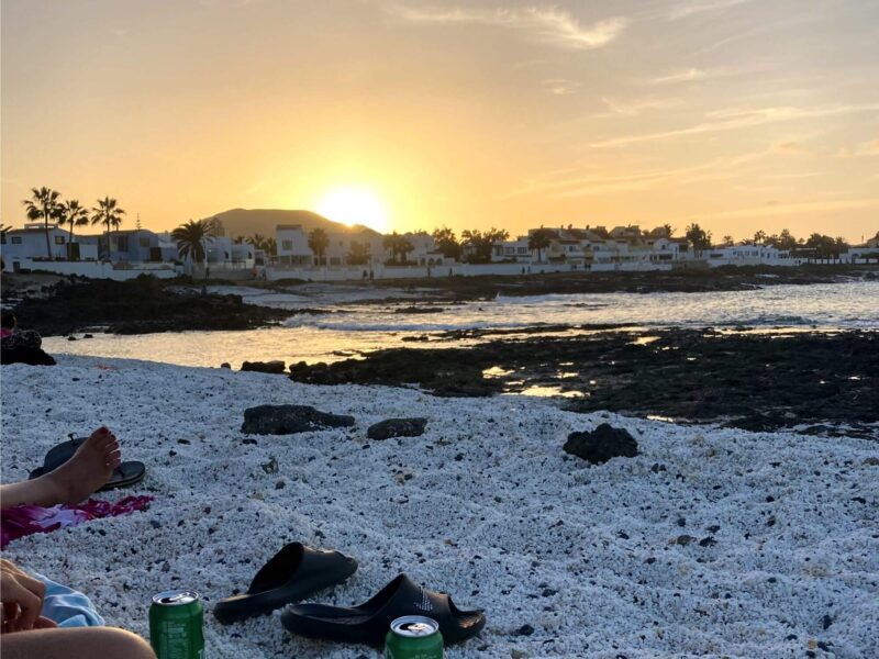 Das hochgeladene Bild zeigt einen friedlichen Strand bei Sonnenuntergang, der eine entspannte "Fuerteventura Party"-Atmosphäre vermittelt. Der Corralejo Strand besteht aus hellem, grobkörnigem Sand oder Kieselsteinen. Im Vordergrund sind persönliche Gegenstände wie Sandalen und eine Getränkedose zu sehen, während Menschen am Strand entspannen. Felsige Bereiche führen ins ruhige Meer, das die goldenen Farben des Sonnenuntergangs reflektiert. Im Hintergrund stehen weiße Häuser einer Küstensiedlung, umgeben von Palmen. Die Sonne geht hinter den Häusern und einem Hügel am Horizont unter, der Himmel ist in goldene und orangefarbene Töne getaucht, was der Szene eine warme und gemütliche Atmosphäre verleiht. Das Corralejo Nachtleben kann mit dem Sonnenuntergang beginnen.