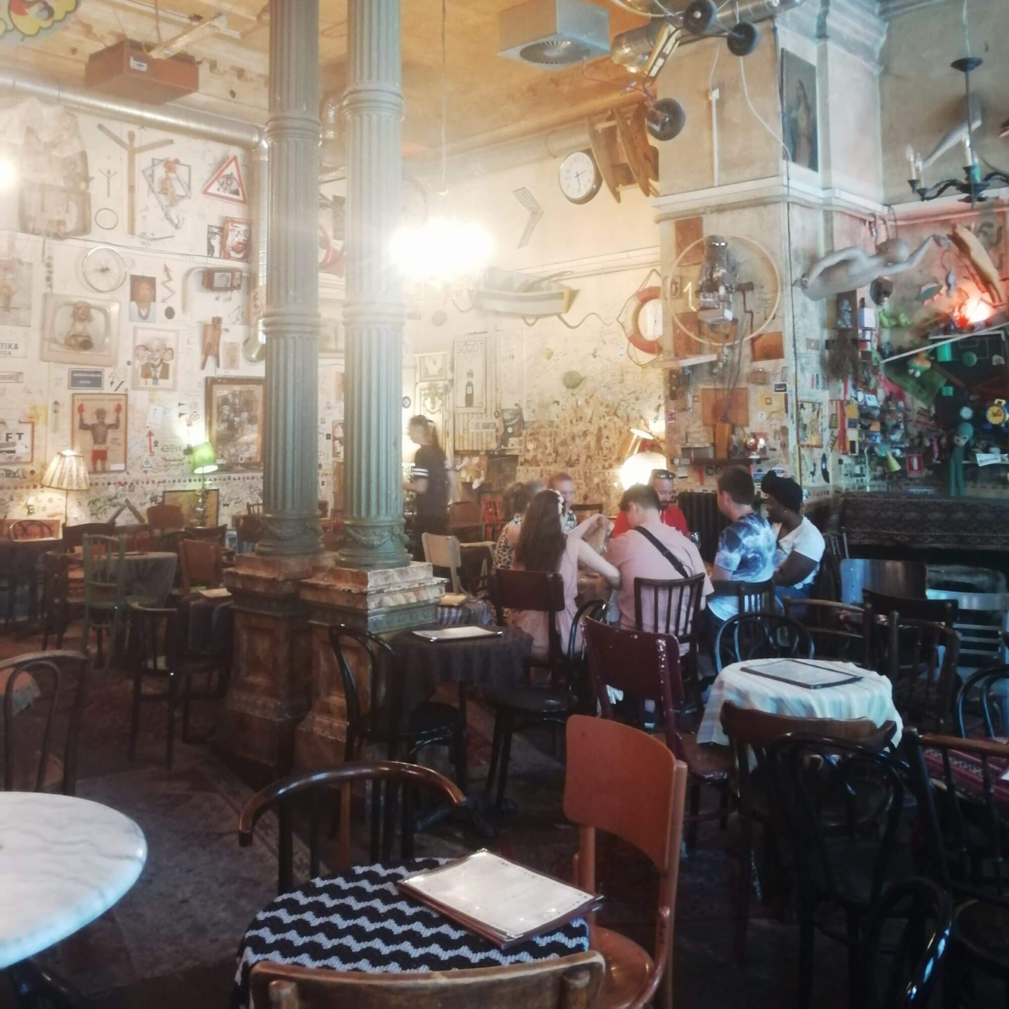 The picture shows the colourful and eclectically furnished bar Csendes, which exudes the typical charm of a ruin bar in Budapest. The bar has a high ceiling on which various decorative objects such as bicycles, clocks and lamps are mounted. Two large, ornate columns in the centre of the room add to the historical atmosphere. The walls are covered with a colourful mix of posters, pictures, signs and old objects, giving the room a creative and slightly chaotic feel. The furniture consists of a variety of tables and chairs, all different and seemingly randomly arranged, which perfectly captures those typical look. In the centre of the picture you can see a group of people sitting at a table chatting. The lighting is subdued, with warm light sources creating a cosy and inviting atmosphere. A waiter or waitress can be seen in the background attending to the guests.