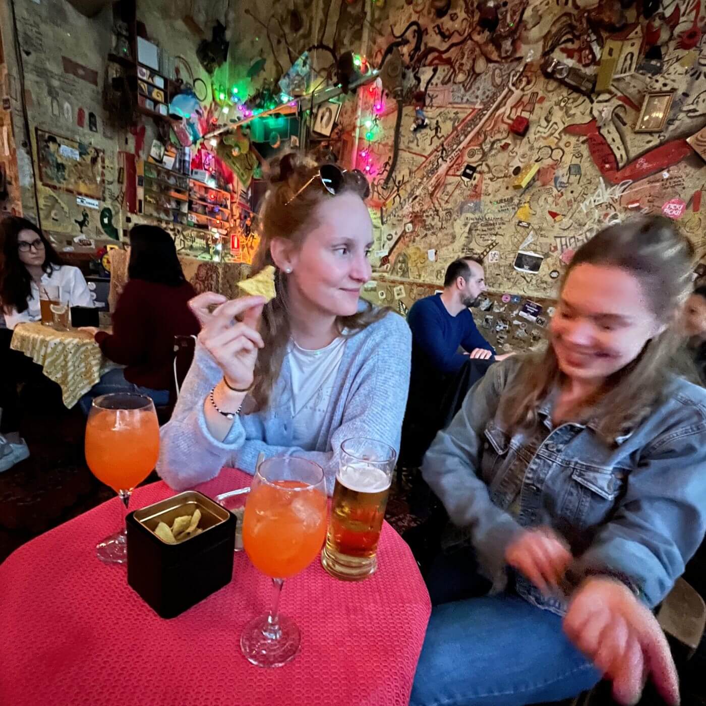 The picture shows a lively scene in one of the famous Ruin Bars Budapest. The interior is decorated with a chaotic but charming assortment of graffiti, drawings, and various items on the walls, with colorful string lights adding to the vibrant atmosphere. In the foreground, two women sit at a table with a red tablecloth. The woman on the left holds a tortilla chip and smiles at her friend, while the woman on the right, slightly blurred, looks down and smiles. On their table are two glasses of orange drinks with ice, a beer glass, and a small black container with tortilla chips. The cozy and convivial atmosphere captures people enjoying their drinks and each other's company in a uniquely decorated ruin bar in Budapest.