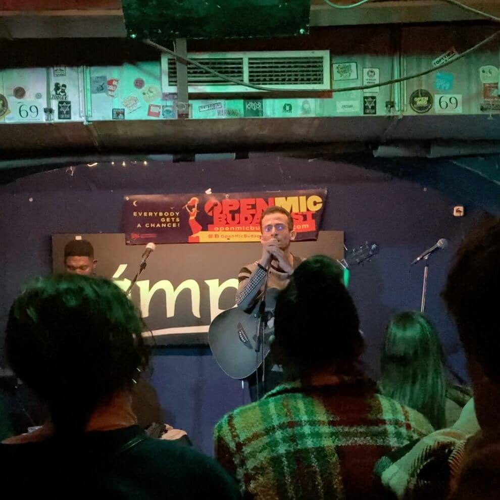 The picture shows a scene in the Lámpás Bar in Budapest. In the foreground you can see several spectators focussing on a stage. On the stage is a musician with a guitar and a microphone, who is speaking or singing. Next to him is another musician with a microphone. The stage has a simple design, with a dark blue background and a sign that reads "Open Mic Budapest". Various stickers and posters can be seen above the stage, creating a creative and relaxed atmosphere. The lighting is subdued, which contributes to the cosy and intimate atmosphere of the bar. The audience are in casual dress and appear relaxed and attentive, indicating a pleasant and friendly atmosphere. Overall, the image gives an impression of a lively and charming underground bar that is a favourite meeting place for music lovers and friends of alternative Budapest".