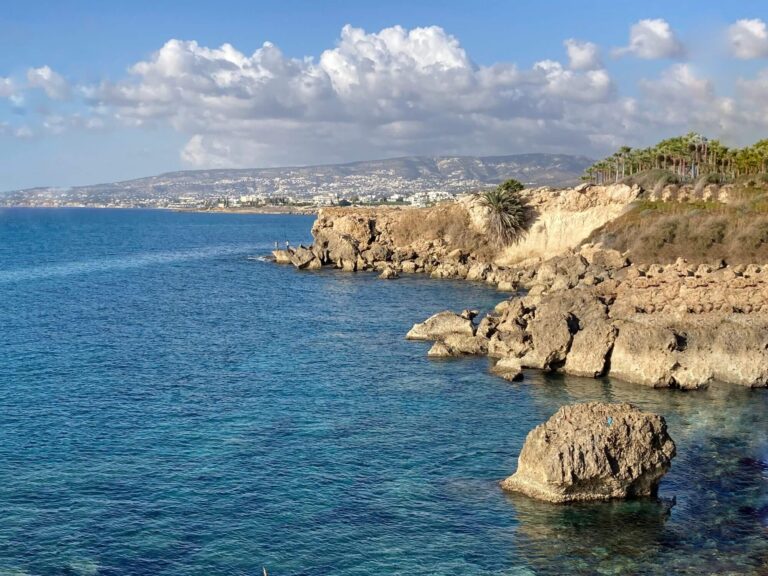 The picture shows a picturesque coastal landscape in Cyprus. In the foreground, the clear, turquoise-coloured waters of the Mediterranean wash around the rocks and stones along the coast. The surface of the water is calm and reveals the rocks below, making the picture appear particularly lively. The coast of Paphos, Cyprus is characterised by rugged, brown rock formations that extend into the sea. Some of the larger rocks protrude strikingly from the water and create a wild, natural panorama. Lush palm trees can be seen on the right of the picture, creating a tropical atmosphere. In the background, the coastline stretches further and offers a view of the distant mountains. On the horizon you can see the settlement of Paphos with its white buildings, which blend harmoniously into the landscape. The scenery shows why Paphos is ideal if you are wondering where to go on holiday in Cyprus. The combination of crystal clear waters, dramatic rock formations and tropical palm trees provide a perfect example of the diverse landscape and picturesque views Cyprus has to offer.