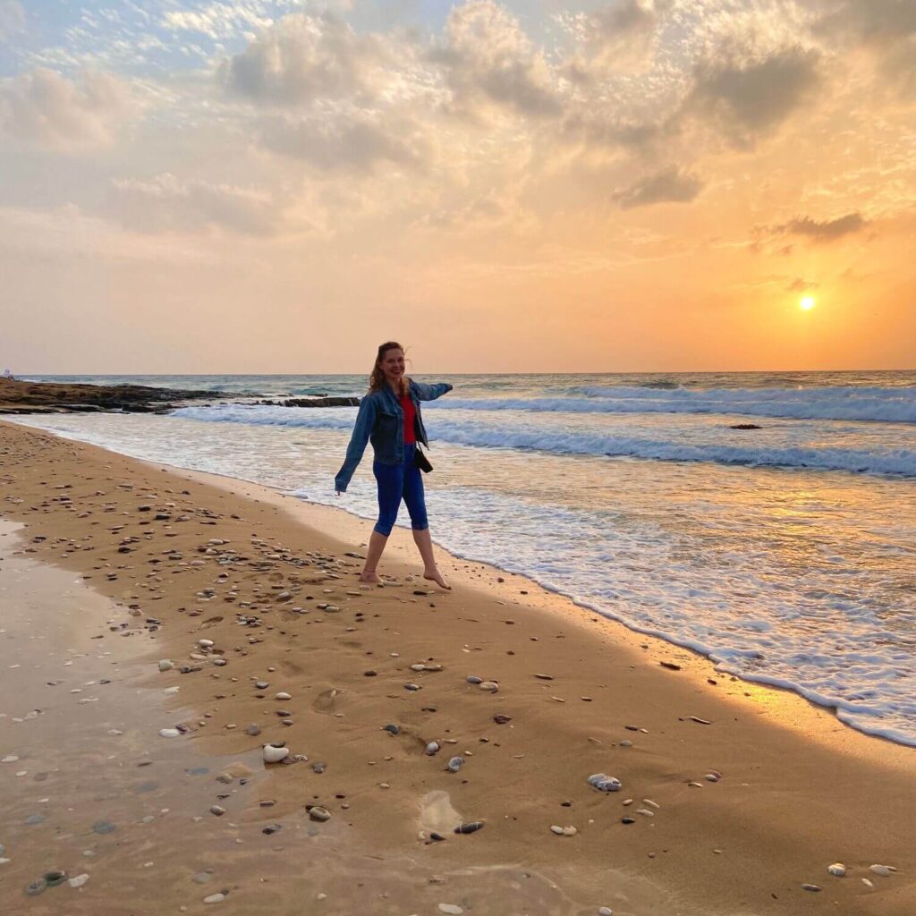 Besonders für die wunderschönen Traumstrände in Zypern. Man sieht mich, eine junge blonde Frau, in langer Jeanshose und Jeansjacke am Sandy Beach Strand herumspazieren. Ich grinse vor lauter glück. Im Hintergrund geht gerade die Sonne über dem Meer unter und färbt den gesamten Himmer orange und rot.