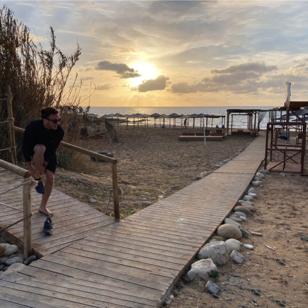 Das Bild zeigt einen idyllischen Strand bei Sonnenuntergang Zypern. Im Vordergrund sieht man einen Mann, der sich auf einem hölzernen Steg seine Schuhe anzieht. Der Steg führt direkt zum Strand, der mit Liegestühlen und Sonnenschirmen ausgestattet ist. Der Himmel ist von einem warmen, goldenen Licht durchzogen, während die Sonne langsam über dem Horizont des Mittelmeers untergeht. Die Wolken sind teilweise von der untergehenden Sonne beleuchtet, was dem Bild eine romantische und friedliche Atmosphäre verleiht. Die Strandlandschaft ist ruhig und einladend, mit einigen wenigen Menschen, die den Sonnenuntergang genießen. Rechts im Bild sind einfache Holzstrukturen zu sehen, die möglicherweise als Strandhütten oder Umkleidekabinen dienen. Dieses Bild fängt perfekt die entspannte Atmosphäre ein und zeigt, warum dieser Ort ideal ist, wenn man sich fragt, wo in Zypern Urlaub machen. Die Kombination aus malerischen Sonnenuntergängen, ruhigen Stränden und einer friedlichen Umgebung macht dies zu einem perfekten Urlaubsziel.