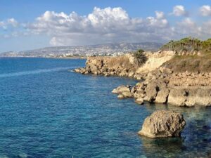 Das Bild zeigt eine malerische Küstenlandschaft in Zypern. Im Vordergrund erstreckt sich das klare, türkisfarbene Wasser des Mittelmeers, das die Felsen und Steine entlang der Küste umspült. Die Wasseroberfläche ist ruhig und lässt die darunterliegenden Felsen erkennen, was das Bild besonders lebendig erscheinen lässt. Die Küste von Paphos, Zypern ist geprägt von schroffen, braunen Felsformationen, die sich bis ins Meer erstrecken. Einige größere Felsen ragen markant aus dem Wasser heraus und schaffen ein wildes, natürliches Panorama. Rechts im Bild sind üppige Palmen zu sehen, die eine tropische Atmosphäre vermitteln. Im Hintergrund zieht sich die Küstenlinie weiter fort und gibt den Blick auf die entfernten Berge frei. Am Horizont sieht man die Siedlung von Paphos mit ihren weißen Gebäuden, die sich harmonisch in die Landschaft einfügen. Die Szenerie zeigt, warum Paphos ideal ist, wenn man sich fragt, wo in Zypern Urlaub machen. Die Kombination aus kristallklarem Wasser, dramatischen Felsformationen und tropischen Palmen bietet ein perfektes Beispiel für die vielfältige Landschaft und die malerischen Ausblicke, die Zypern zu bieten hat.