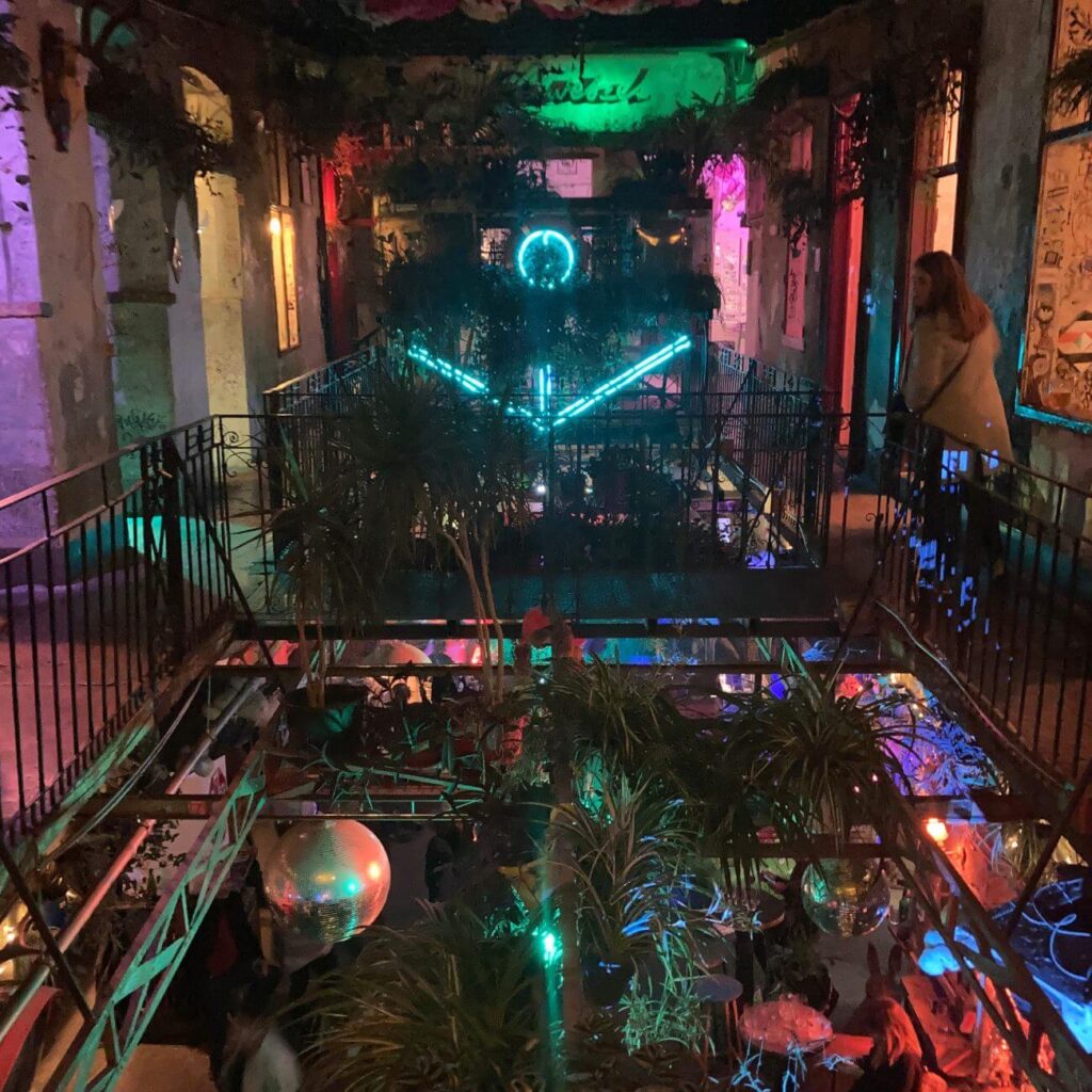 Szimpla Kert bei Nacht. Man ist auf einer Galerie im ersten Stock und sieht hinunter auf Pflanzen, eine Diskokugel und viele bunte Lichter beleuchte das gesamte Gebäude.