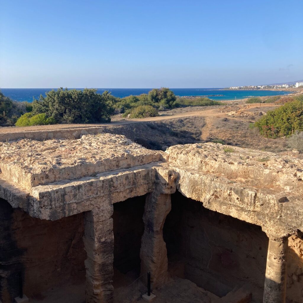 Das Bild zeigt die historische Stätte "Tombs of the Kings" in Paphos. Im Vordergrund sieht man die beeindruckenden Felsengräber, die in den Boden gehauen wurden. Die massiven Steinstrukturen und die aus dem Felsen gehauenen Säulen geben einen Einblick in die antike Architektur. Die Oberfläche der Gräber ist rau und zerklüftet, was die lange Geschichte und die Verwitterung über die Jahrhunderte hinweg widerspiegelt. Im Hintergrund erstreckt sich eine natürliche Landschaft mit dichten Büschen und Bäumen, die von einem gut sichtbaren Pfad durchzogen wird. Der Pfad führt zu einer Küstenlinie, die das Bild in der Ferne dominiert. Das azurblaue Mittelmeer funkelt unter dem klaren Himmel und schafft einen beeindruckenden Kontrast zu den erdigen Tönen der antiken Gräber und der umgebenden Vegetation. Am Horizont erkennt man die Umrisse von Gebäuden und eine kleine Ansammlung von weißen Häusern, die auf eine nahegelegene moderne Siedlung hinweisen. Der Himmel ist klar und wolkenlos, was auf einen typischen sonnigen Tag in Zypern hindeutet. Wenn du dich fragst, wo in Zypern Urlaub machen, ist Paphos mit seinen historischen Stätten und der wunderschönen Küstenlandschaft ein idealer Ort. Die Szenerie vereint die historischen und natürlichen Schönheiten und zeigt, warum dieser Ort ein beliebtes Reiseziel ist.