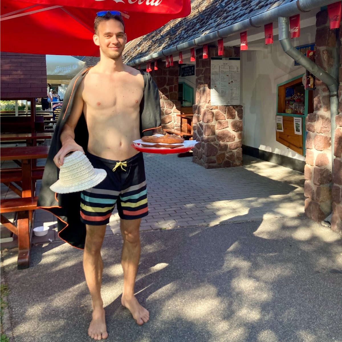  The image features a cheerful young man enjoying his time at Lake Balaton. He's casually dressed in striped swim trunks and a draped towel, holding a straw hat in one hand. In the other hand, he proudly presents a plate of Lángos, a popular Hungarian fried dough snack. Behind him, the setting is lively with a rustic stone building housing the Napsugár Büfé in Kaptalanfüred, shaded by a bright red Coca-Cola umbrella. Picnic tables and the relaxed atmosphere suggest a perfect spot for a delicious bite and a pleasant day by the lake.