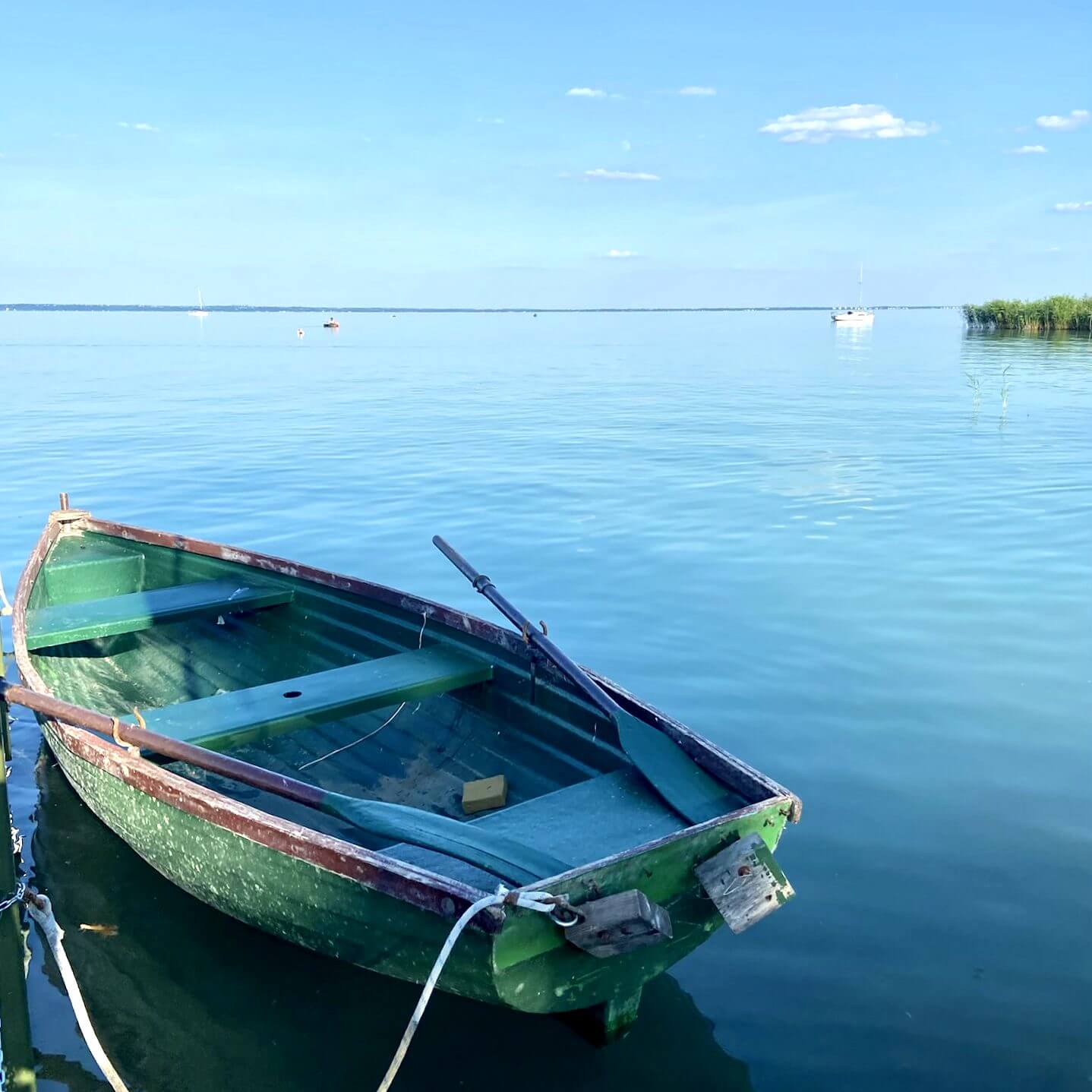 The image showcases a peaceful and serene scene at Lake Balaton, specifically in Kaptalanfüred. A rustic green rowboat is tied to the dock, gently floating on the clear, calm waters of the lake. The tranquil water reflects the blue sky and the few scattered clouds, creating a picturesque and soothing atmosphere. In the distance, a sailboat can be seen, adding to the idyllic charm of the scene. The shoreline, with its lush greenery, is visible on the right, enhancing the natural beauty of the setting. This image perfectly captures the calm and inviting ambiance of a day by Lake Balaton.