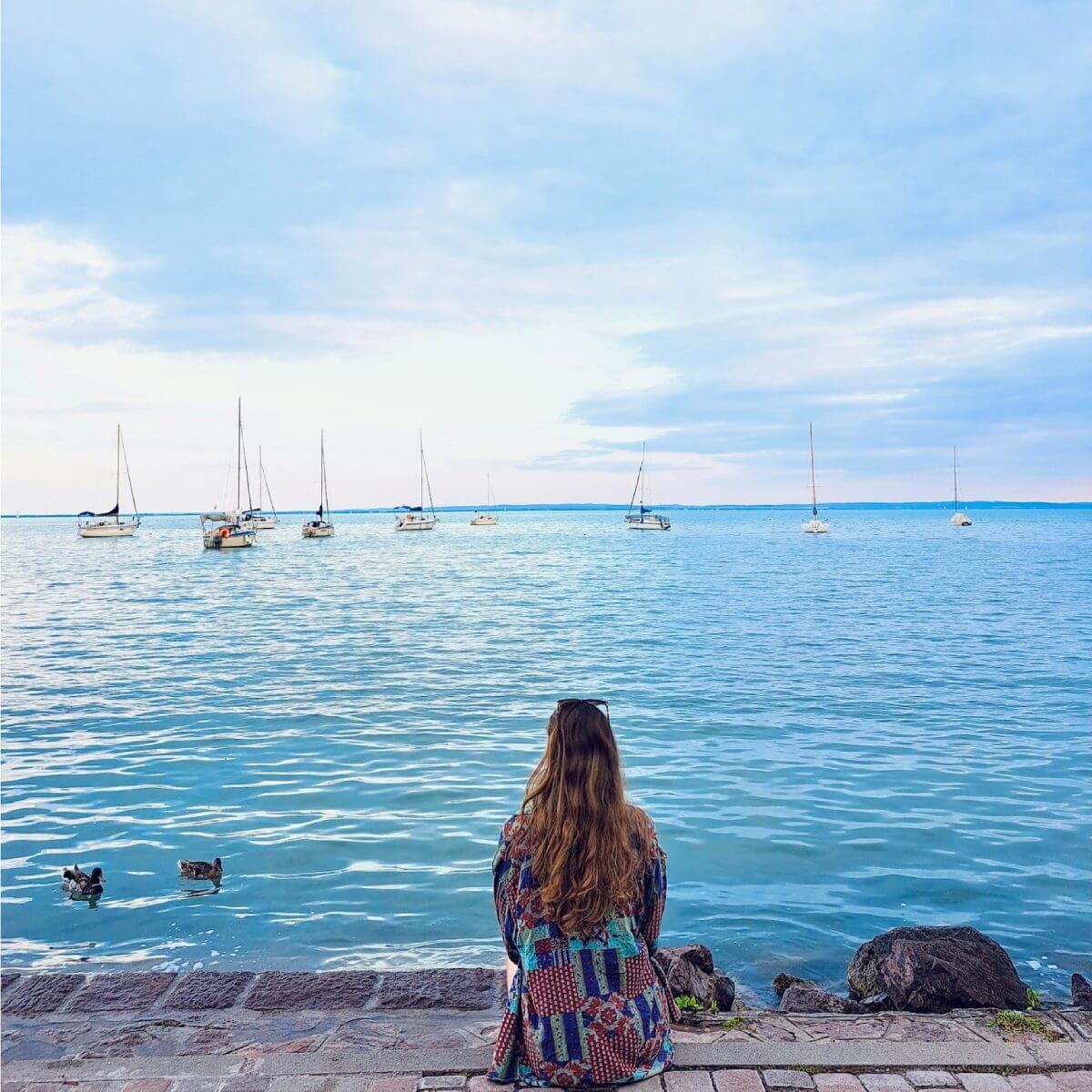 The picture shows the serene view of Lake Balaton next to Balatonalmádi. In the foreground, you can see a woman with long, wavy hair, sitting on a stone-paved edge, facing the lake. She is wearing a colorful, patterned jacket and appears to be enjoying the tranquil scenery. The calm water of the lake stretches out before her, with several sailboats anchored peacefully in the distance. The sky above is partly cloudy, creating a soft, diffused light that enhances the peaceful ambiance of the scene. The overall atmosphere is calm and picturesque, capturing a perfect moment of relaxation by the lake.