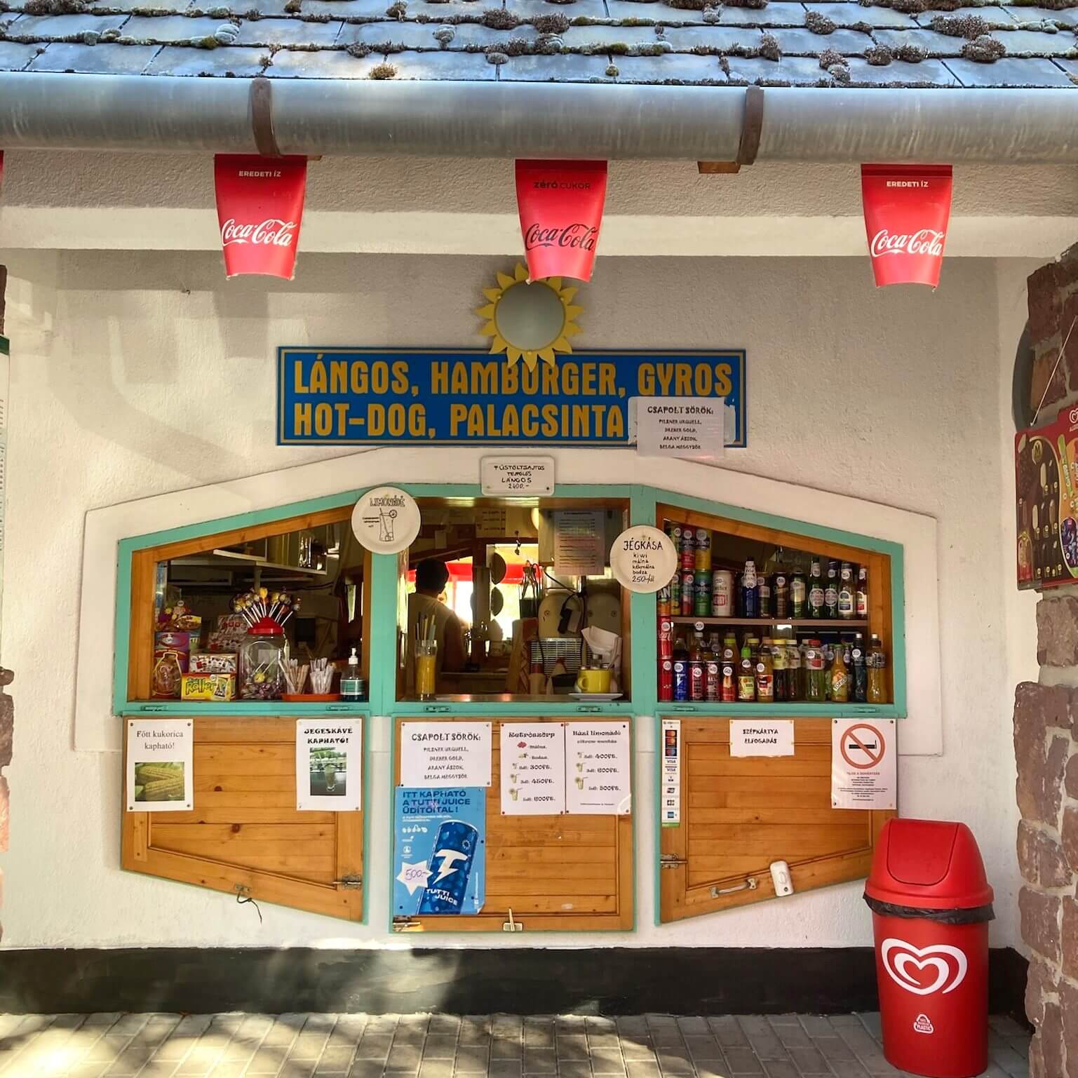 This picture shows a small food stand at the Balaton lake area, likely in Káptalanfüred. The stand, named "Napsugár Büfé," has a charming, rustic appearance with a tiled roof and a bright blue and yellow sign advertising popular Hungarian and fast food items such as lángos, hamburgers, gyros, hot dogs, and palacsinta (pancakes). The stand has a large window through which you can see various snacks, drinks, and a glimpse of the interior where the food is prepared. Coca-Cola banners hang from the roof, and there are several signs and menus displayed around the window, listing prices and available items. A red trash bin with an ice cream logo stands to the right, indicating that ice cream might also be available at the stand. The overall atmosphere is inviting and casual, typical of a local eatery in a popular tourist area by the lake.