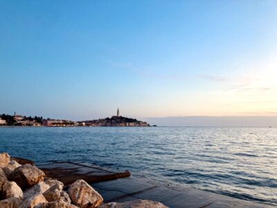 View of the historic town of Rovinj, Croatia, at sunset from across the calm Adriatic Sea. The iconic bell tower of St. Euphemia’s Church rises above the picturesque old town, which is illuminated with soft lights. In the foreground, large rocks and a concrete platform border the peaceful water, enhancing the tranquil coastal atmosphere. The sky transitions from blue to a gentle golden hue, perfectly capturing the serene beauty of Rovinj Croatia.