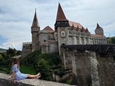 Frau sitzt in einem blauen sommerkleid vor der großen Burg Corvinus in Rumänien Transsilvanien.