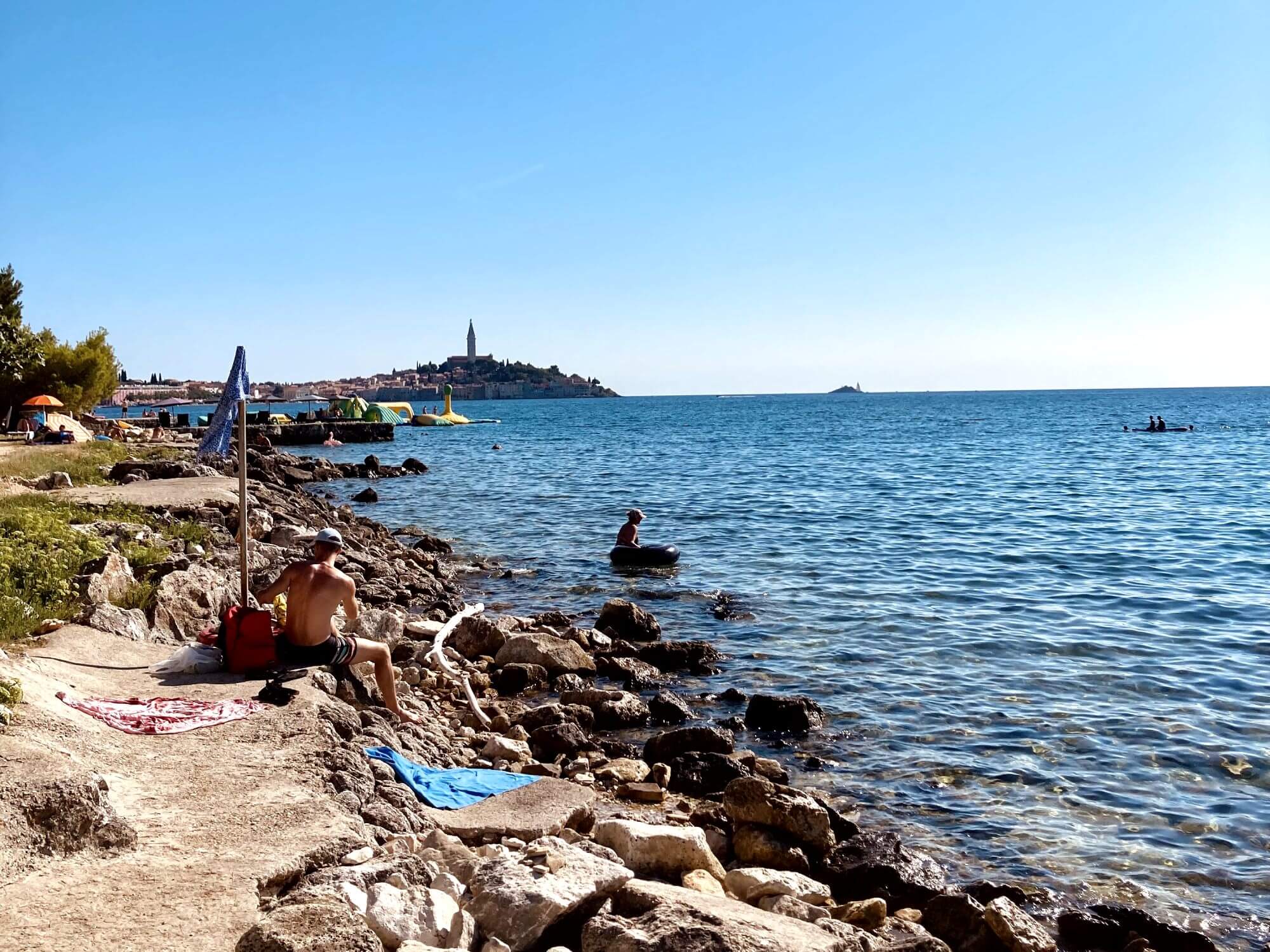 In this picture you can see the cosy Rovinj beach called Sand Beach Biondi. Some people are lolling in the sun and in the background you can see the old town of rovinj. the sea stretches out to the right of the picture. It is a sunny day.