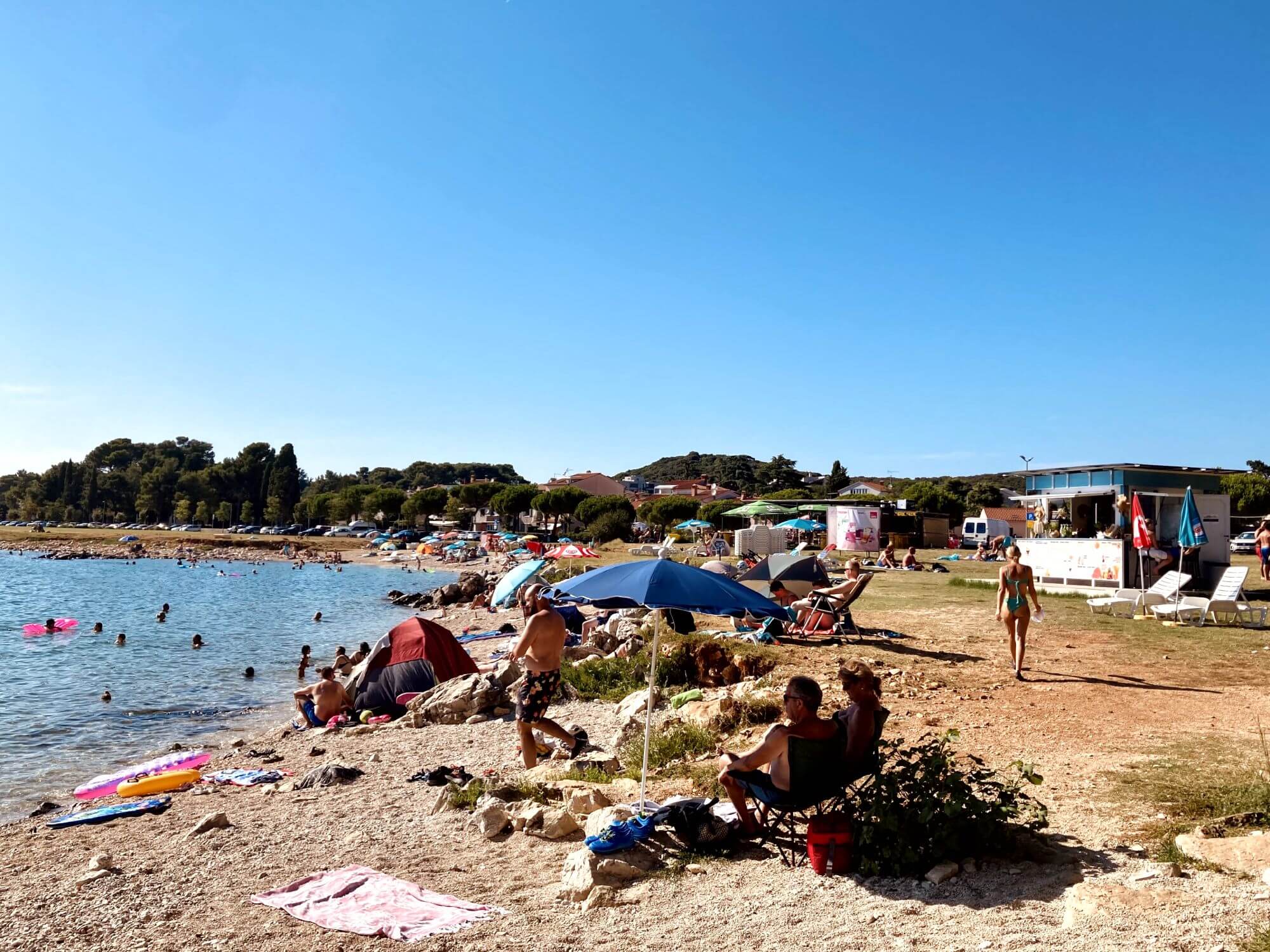 In this picture you can see the Rovinj beach called Sand Beach Biondi. Some people are lolling in the sun and in the background you can see two beach bars with the delicious aperol Spritz. You can also see a forest with pine trees in the background. People enjoying the sunny day.