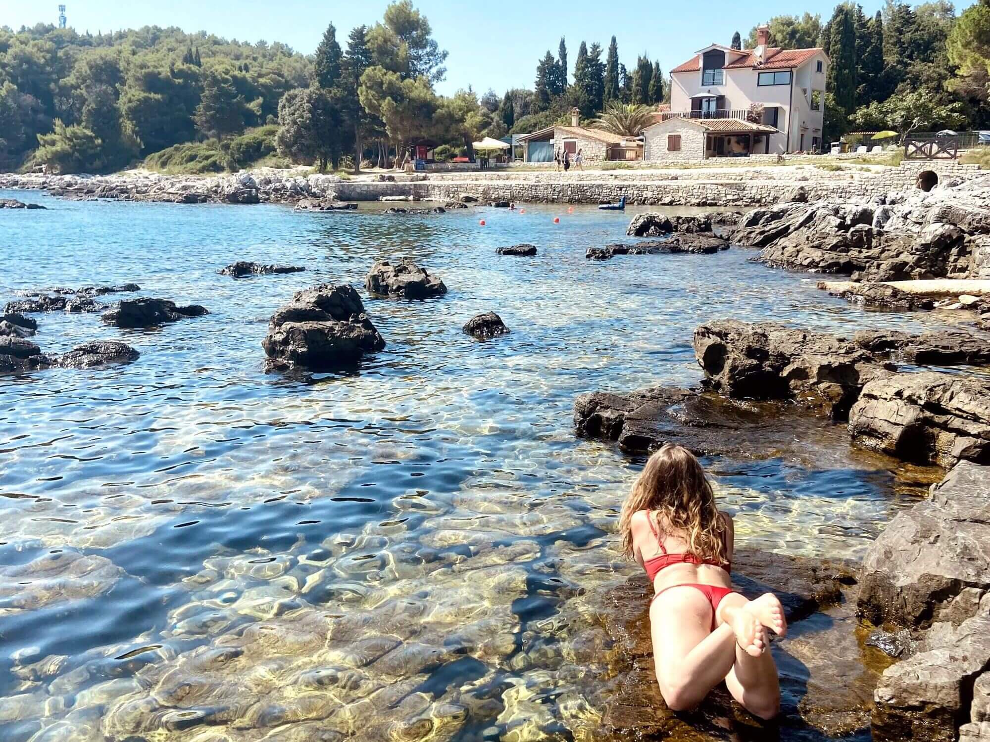 This picture of Grote Beach shows a woman lying on a flat rock in shallow, clear water, wearing a red bikini, her feet crossed as she relaxes. The Adriatic Sea is crystal-clear, revealing rocks beneath the surface, with larger rocks scattered along the shoreline. In the background, lush greenery covers a hillside, and a few charming houses sit near the coast. The scene is peaceful, showcasing the natural beauty of one of the more secluded Rovinj beaches in Rovinj Croatia.