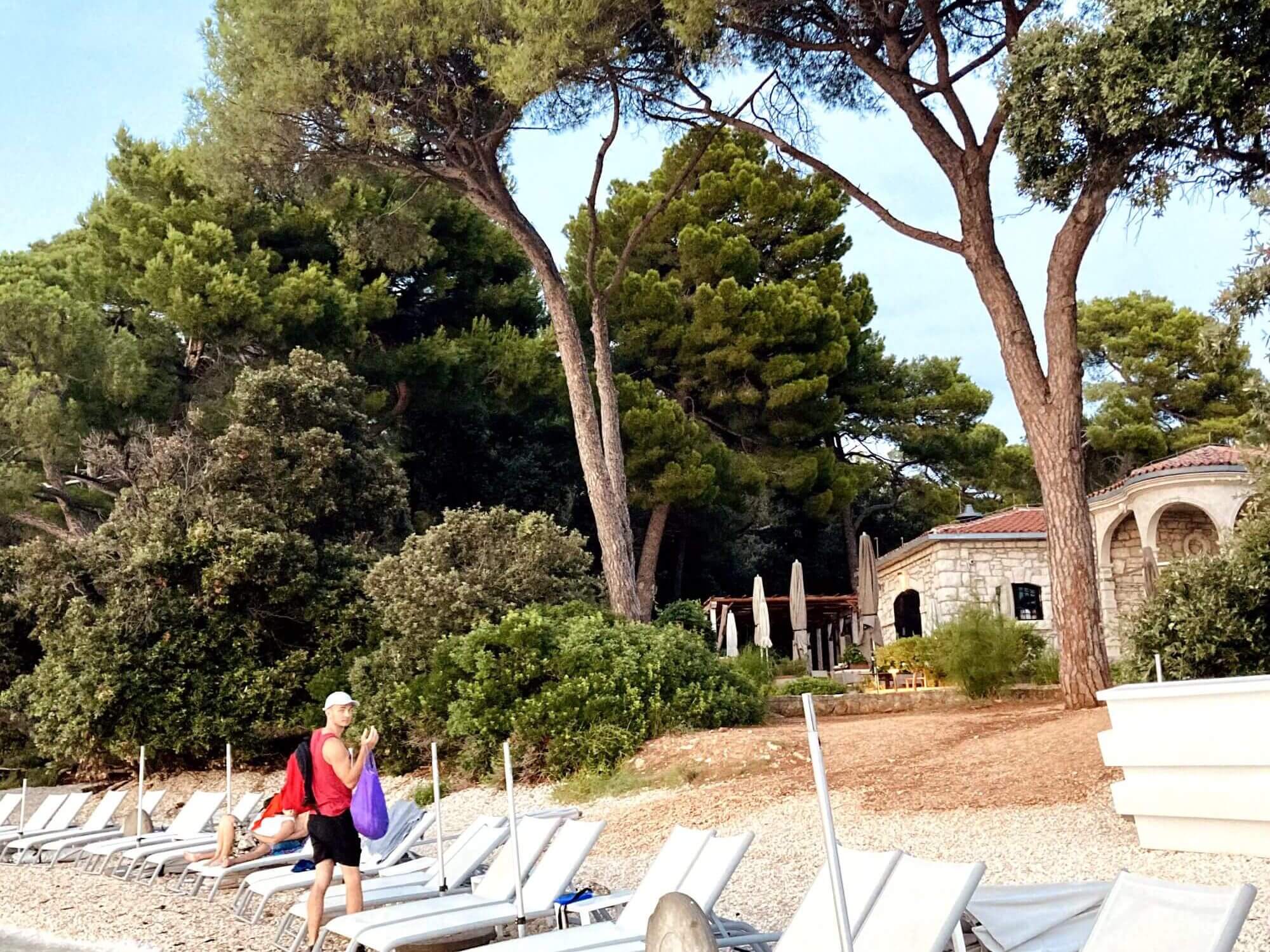 The picture shows a quiet beach in Rovinj, near the Grill Lovor restaurant. In the foreground, empty sun loungers lie on the pebble beach while a man with a bag walks relaxed along the shore. Tall pine trees, typical of forest beaches in Croatia, surround the scene. In the background is the charming stone building of Grill Lovor, which exudes Mediterranean flair. This idyllic combination of nature and relaxation perfectly reflects a holiday in Rovinj, where the beach at Grill Lovor offers an oasis of relaxation. 