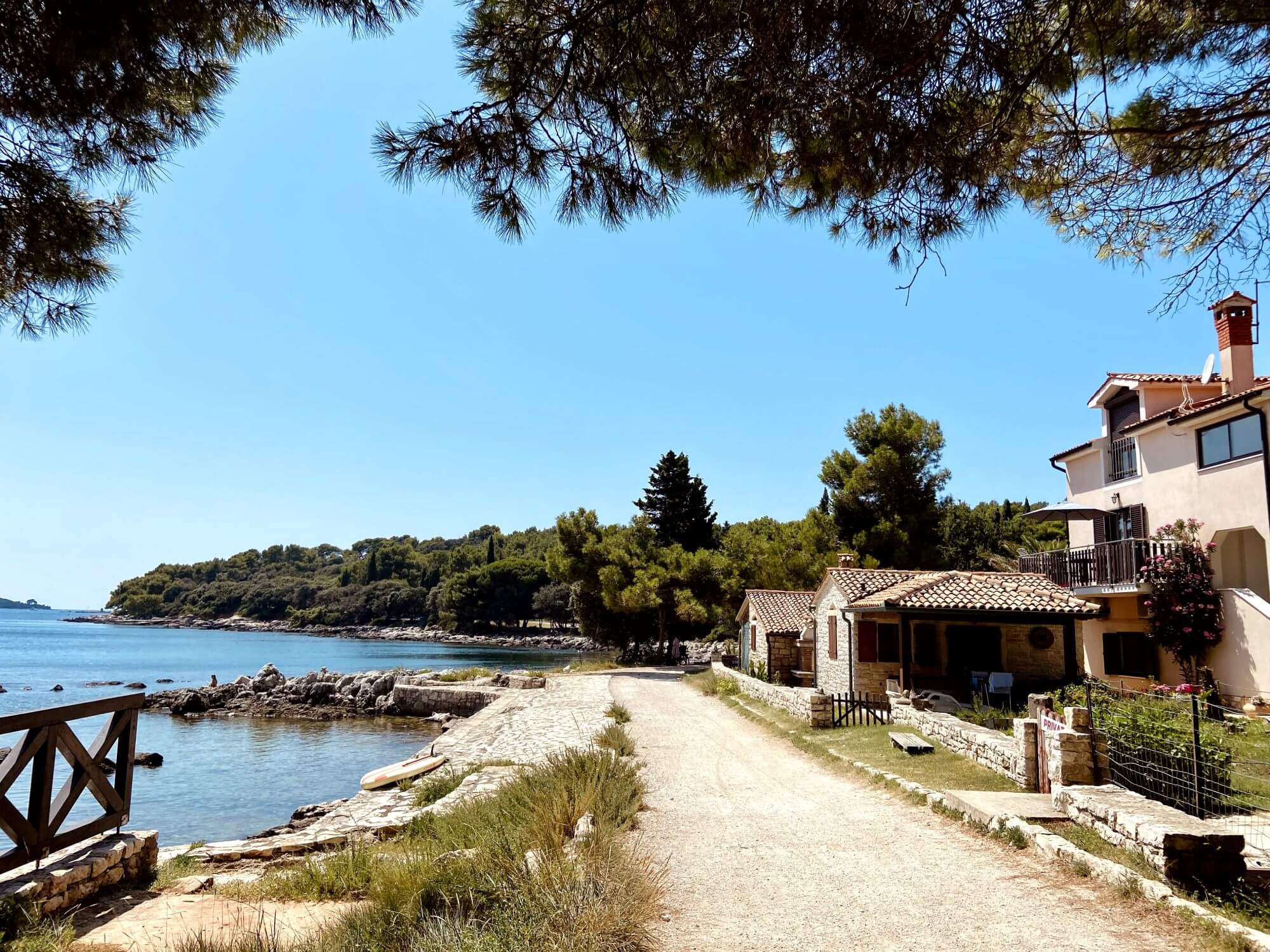 This picture from Grote Beach captures a quiet, scenic path that runs alongside the Adriatic Sea. On one side, the clear blue waters lap against rocky edges, while the other side features charming stone houses with red-tiled roofs, some with balconies overlooking the coast. Tall pine trees frame the scene, casting soft shade onto the path. The distant hillside is covered in lush greenery, with a few people relaxing near the shore. This tranquil spot is a perfect example of the peaceful beauty found on Rovinj beaches, offering a relaxing getaway in Rovinj Croatia.