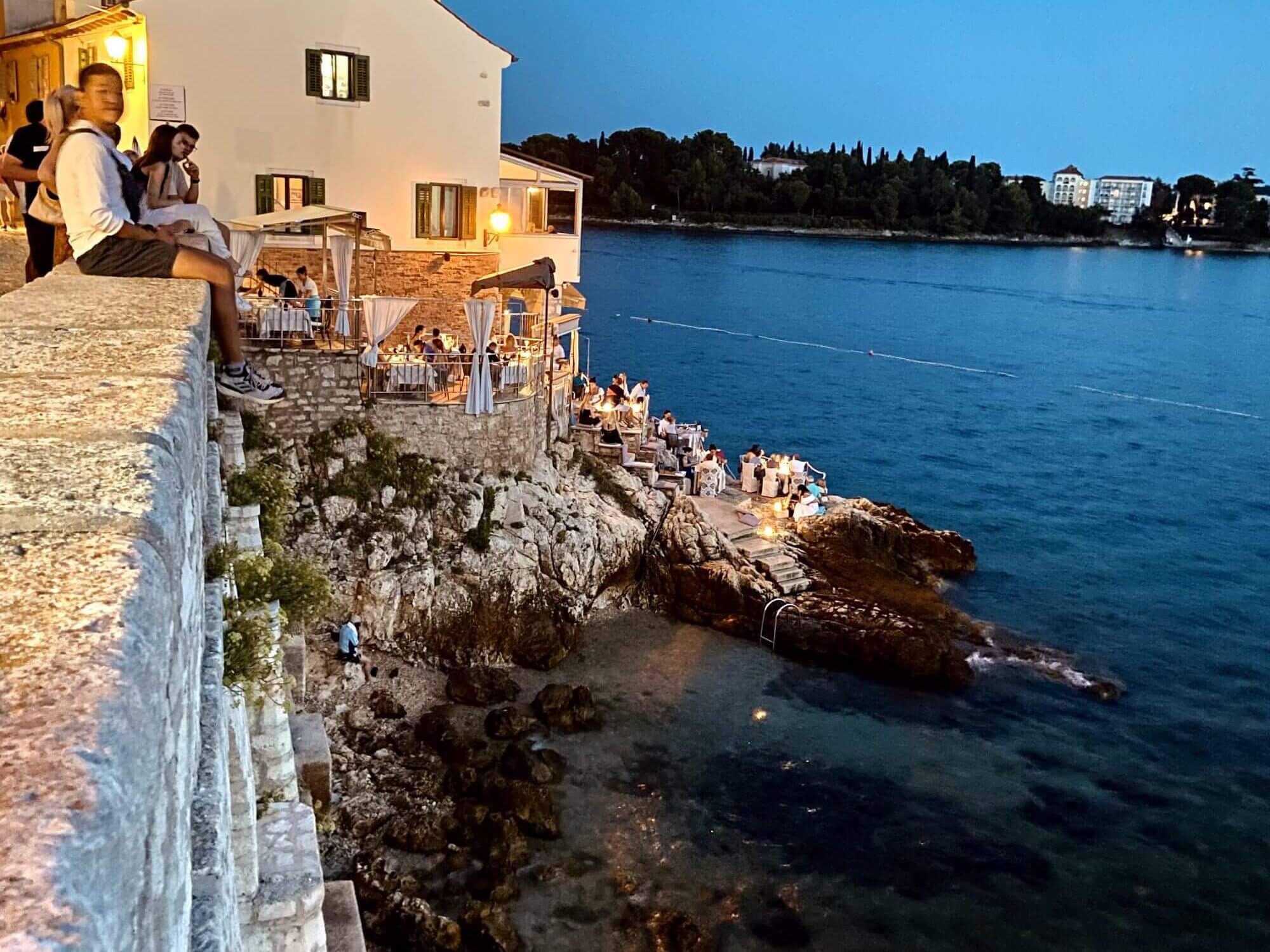 In this picture it is already evening. You can see the rocky town beach of Rovinj, looking down from the wall to the right. Occasionally you can see the rocks protruding from the water. Where it is a little shallower, you can even see the sandy seabed. At the bottom, right next to the beach, you can walk up a staircase to a bar, where guests are still sitting. the whole picture conveys the perfect vibe of Rovinj Croatia. 