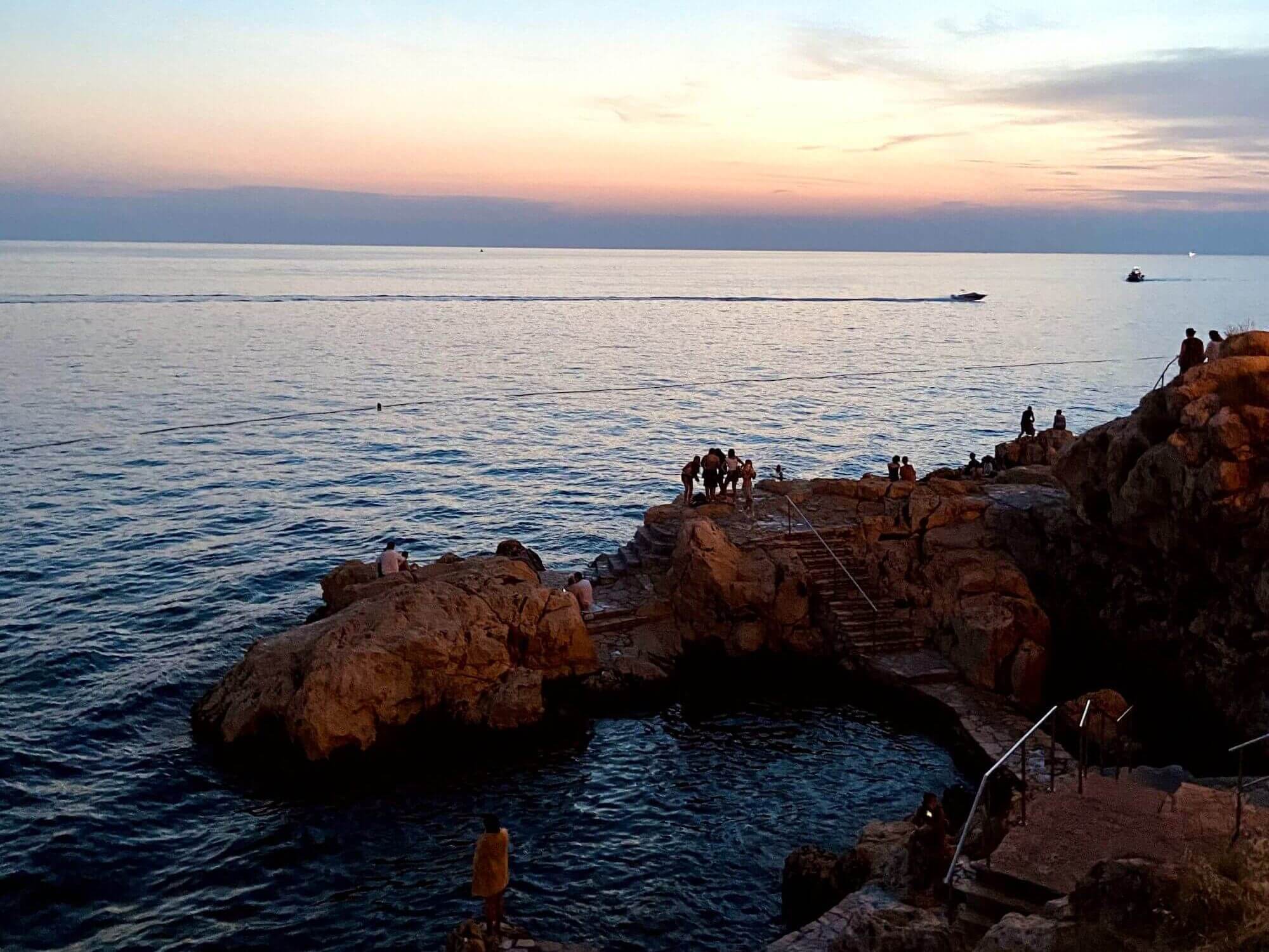 In this picture it is already evening. You can see the rocky town beach of Rovinj if you look down from the wall on the left. Occasionally you can see the rocks sticking out of the water. In the background you can see how the sun has just set. The sky is still reddish and purple in colour. The whole picture reflects the perfect mood of Rovinj Croatia. 