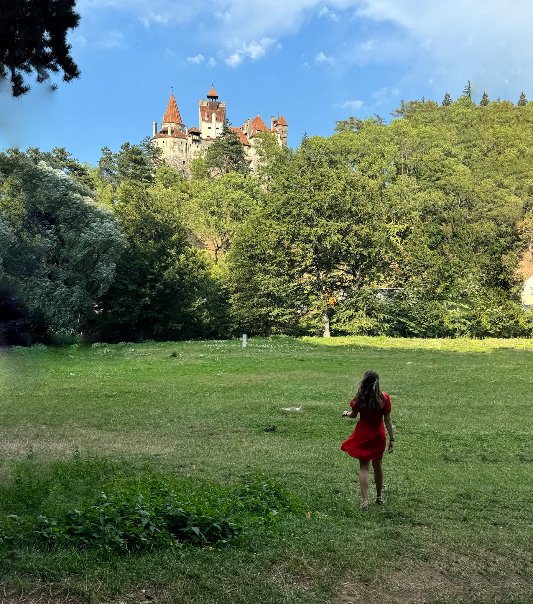 In the distance, behind the trees, you can see the real Dracula Castle. In front, a woman in a red dress strolls through Transylvania, Romania. This location is a must-visit on any Transylvania road trip.