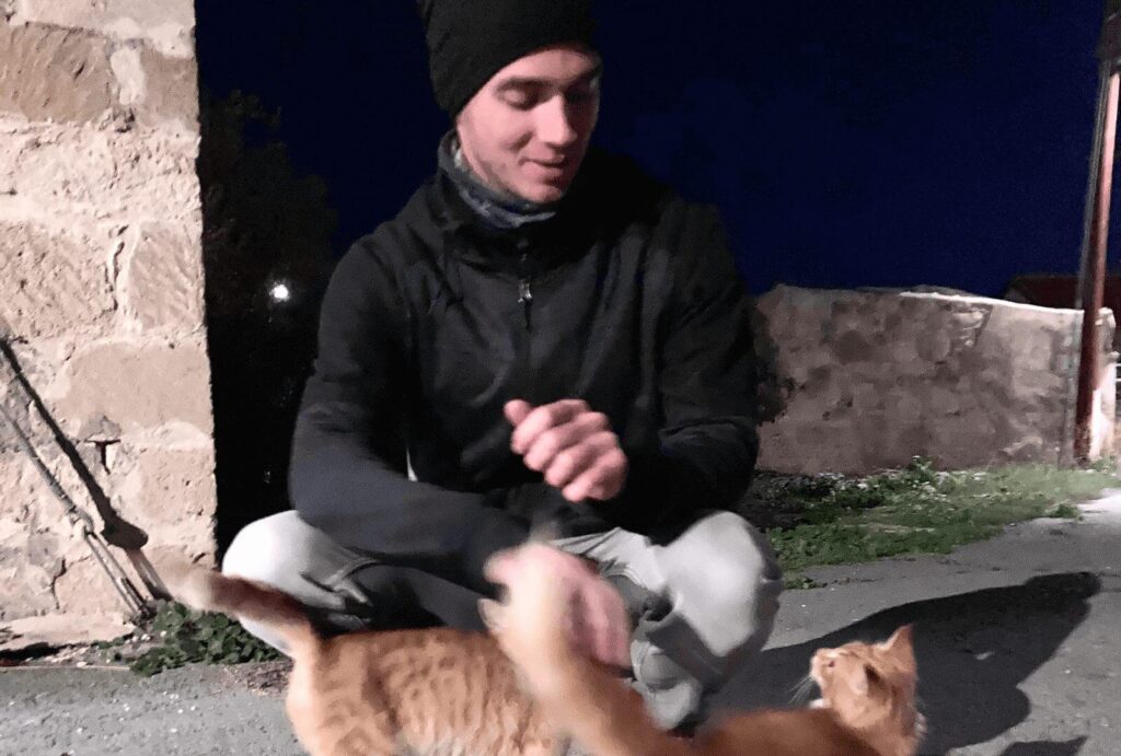 A tall man crouches next to two orange cats from Cyprus in a village with stone houses, petting them. It's nighttime.