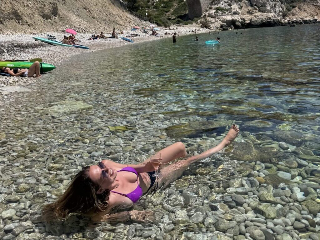 Kristallklares Wasser, weiße Kieselsteine und ein imposantes Aquädukt im Hintergrund – dieses Bild zeigt die entspannte Seite eines Urlaub gegen Hand in Frankreich. Die Frau genießt die Sonne im seichten Wasser einer traumhaften Bucht an der Mittelmeerküste. Solche Plätze laden nicht nur zum Baden und Entspannen ein, sondern sind auch perfekt für alle, die Housesitting am Meer nutzen möchten, um kostenlos zu wohnen und gleichzeitig einen authentischen Einblick in das Leben an Frankreichs Küsten zu bekommen. Zwischen steilen Felsen, türkisfarbenem Meer und mediterraner Atmosphäre lässt sich hier der Sommer auf ganz besondere Weise erleben.