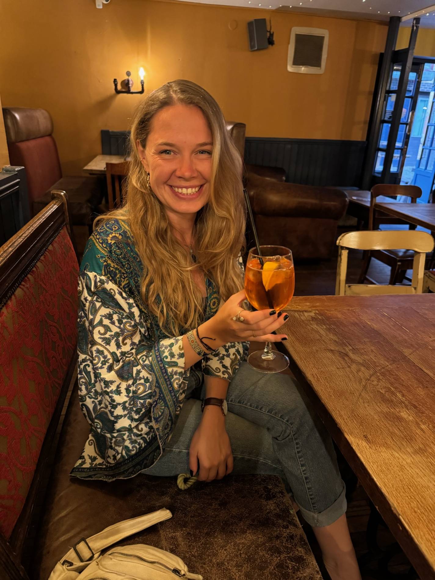 Auf dem Bild sieht man eine Blondine ihren Aperol Spritz in einem Pub in Cambridge genießen. Sie ist mit ihrem Urlaub gegen Hand sehr zufrieden, das erkennt man an ihrem Lächeln.