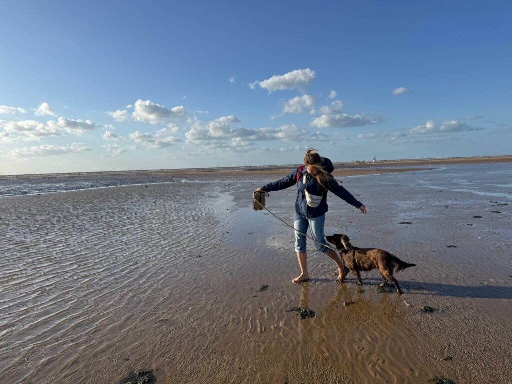 Das Bild zeigt das Thema Was ist Urlaub gegen Hand. Man sieht eine Frau die voller Freude mit einem Cocker Spaniel an einem englischen Strand spazieren geht.