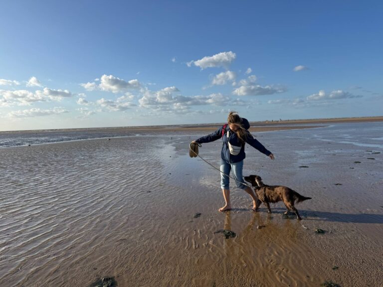 Das Bild zeigt das Thema Was ist Urlaub gegen Hand. Man sieht eine Frau die voller Freude mit einem Cocker Spaniel an einem englischen Strand spazieren geht.