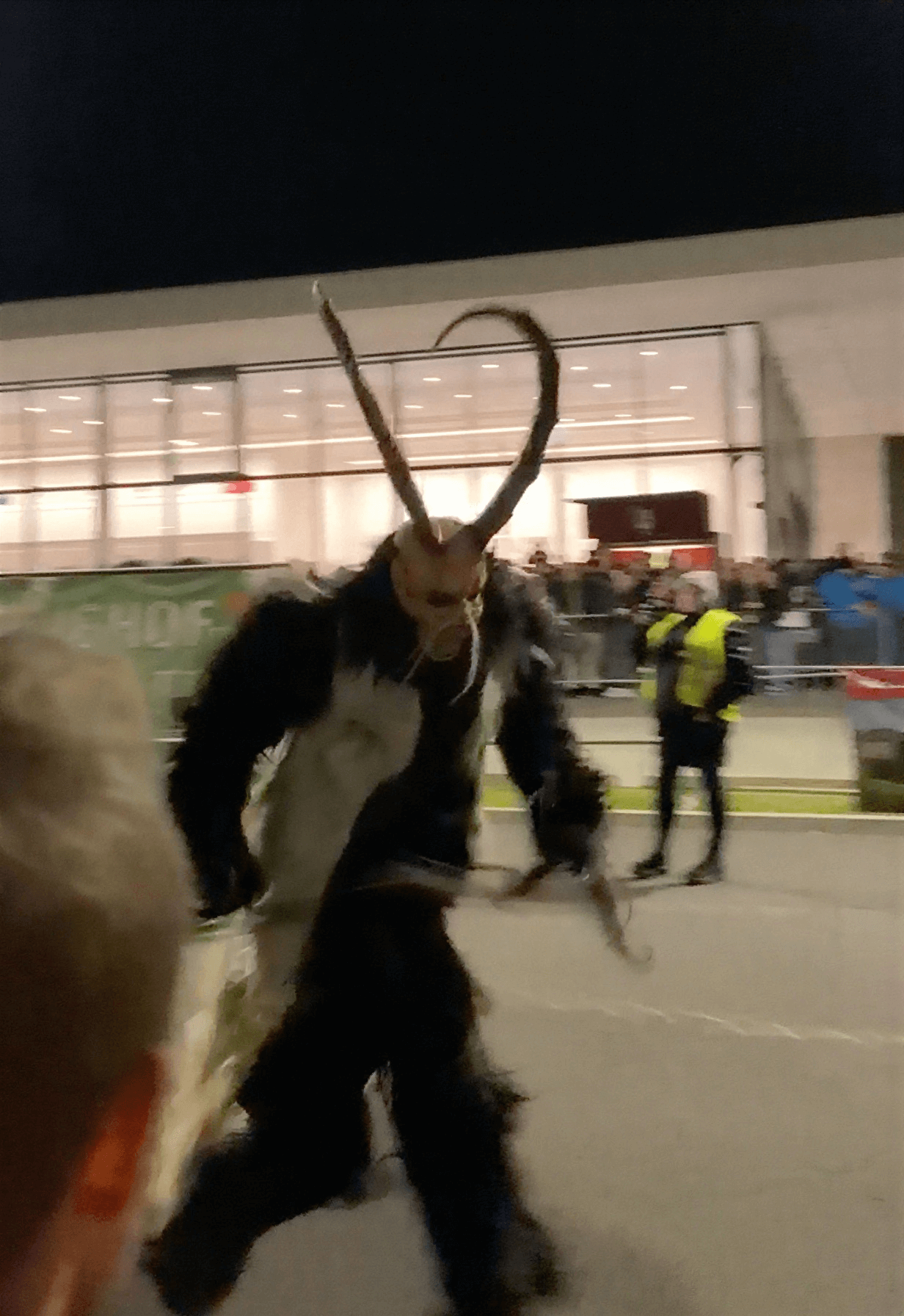 Das Bild zeigt einen Krampus in voller Bewegung. Mit besonders hohen Hörnern und einem schwarz-weißen Fellkostüm läuft er an einer Absperrung vorbei, während Zuschauer und Sicherheitskräfte das Spektakel beobachten. Die dynamische Szene ist charakteristisch für viele Krampus- und Perchtenläufe in Österreich, bei denen die Darsteller mit rasselnden Ketten und lauten Glocken für Atmosphäre sorgen.