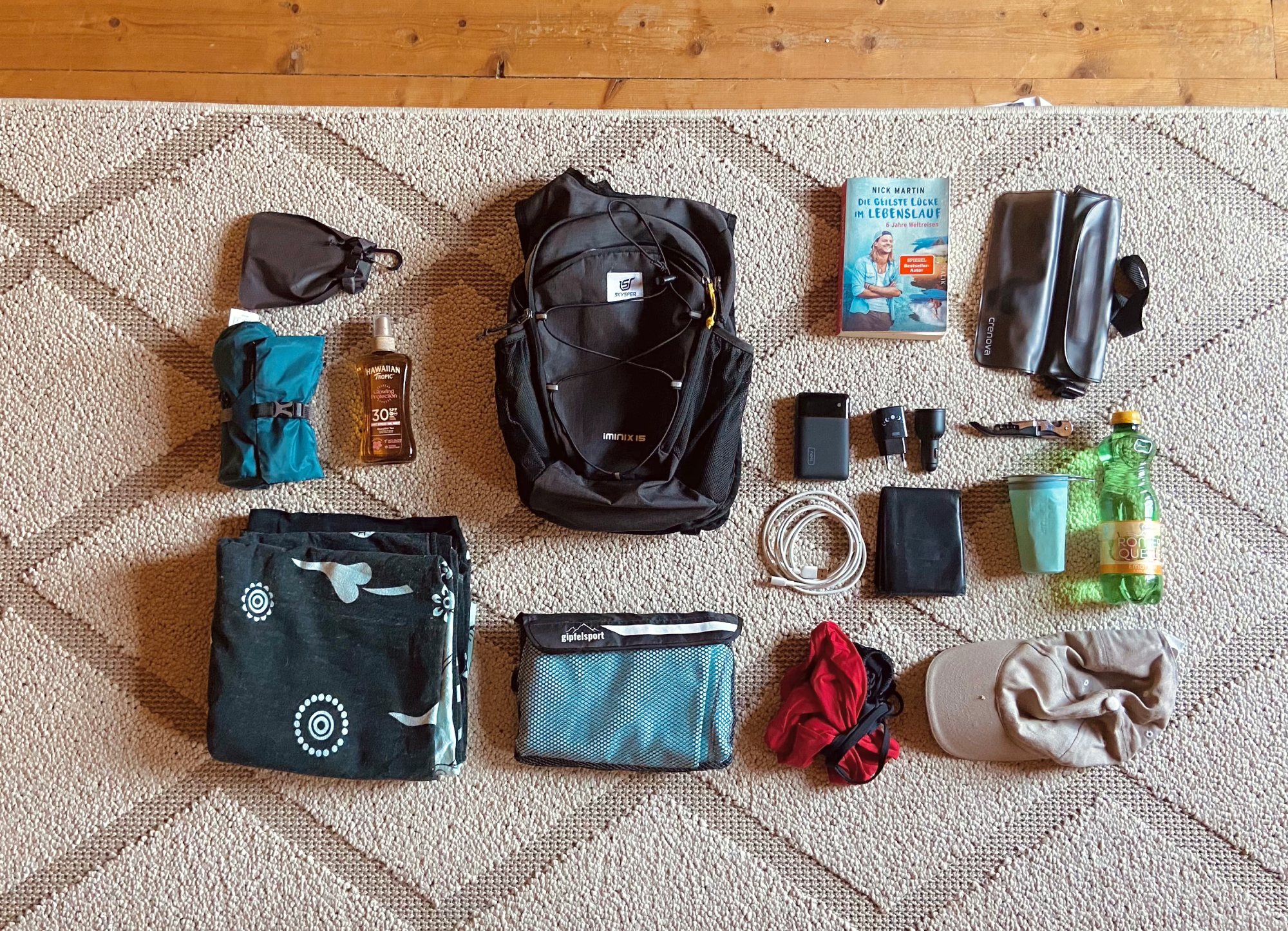 Auf diesem Bild sehen wir einen leeren Tagesrucksack und rundherum leigen alle produkte dich ich darein gepackt habe. Eine schwarze stranddecke, ein mikrofasertuch, und vieles mehr