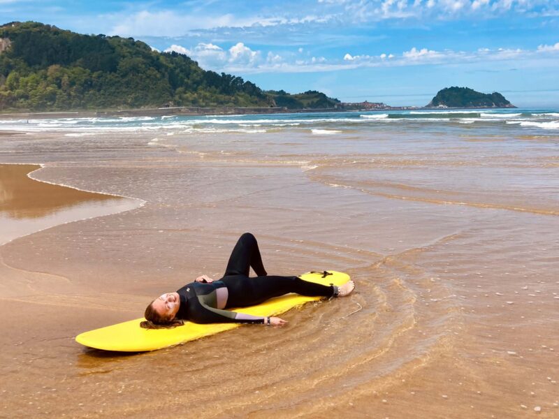 Hier sind man eine surferin die am Strand von Zarautz auf ihrem weichen Softboard liegt. Sie probiert gerade surfen in Spanien aus und hat großes Grinsen im Gesicht.